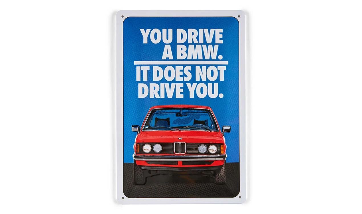 Targa in metallo BMW Lifestyle Primavera/Estate 2025, blu/rosso/bianco, dimensioni 280x200 mm, con il testo "You drive a BMW. It not drive you." e modello E21. Valido per BMW. BMW originale.