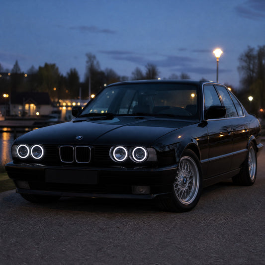 BMW Serie 5 E34 berlina nera all'imbrunire con angel eyes ad anelli LED bianchi nei suoi quattro fari