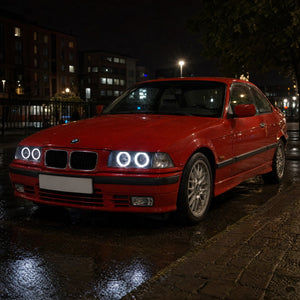 BMW Série 3 E36 coupé rouge de nuit en ville, avec angel eyes LED blancs autour de ses quatre optiques rondes, deux anneaux par phare