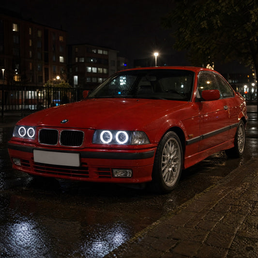 BMW Serie 3 E36 coupé rojo de noche en la ciudad con ojos de ángel LED blancos alrededor de sus cuatro luces redondas, dos aros por faro
