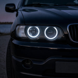 Nahaufnahme des Scheinwerfers eines schwarzen BMW X5 E53 vor dem Facelift in der Abenddämmerung mit zwei leuchtenden weißen LED-Angel-Eyes-Ringen